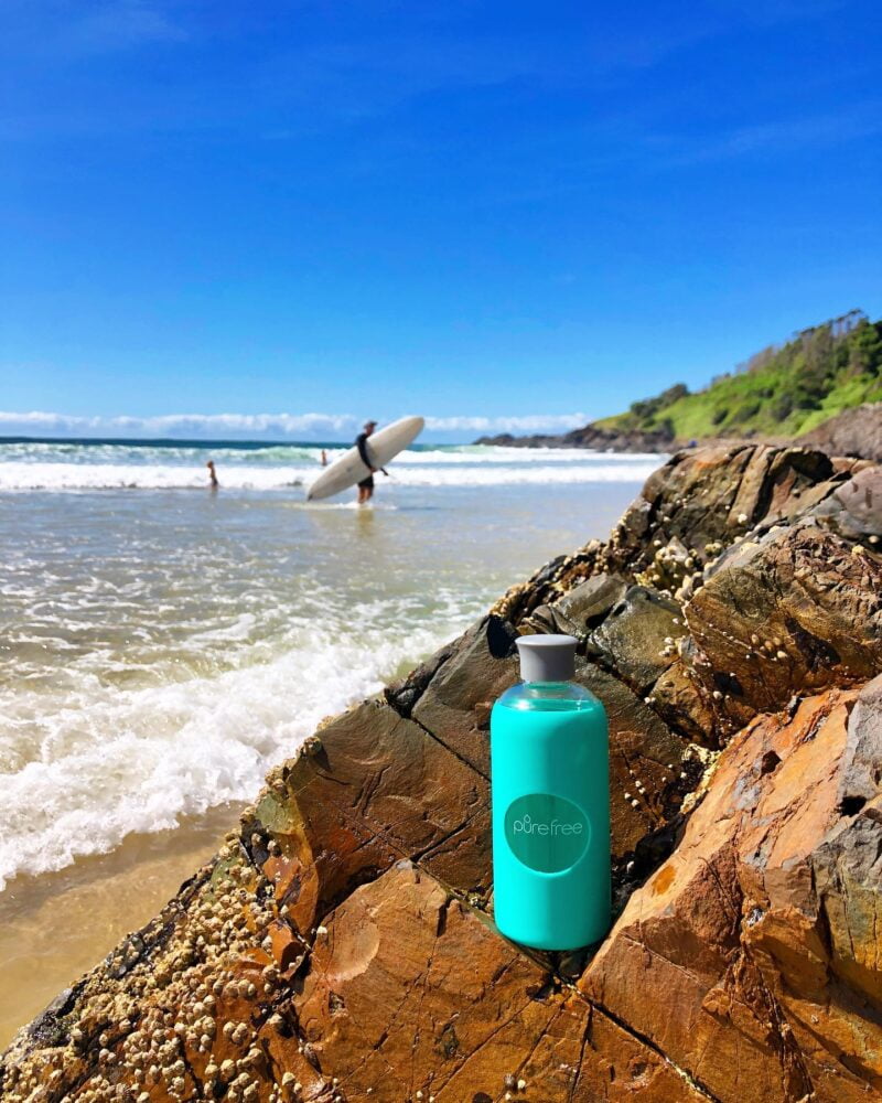 Reusable glass water bottle. 500ml, with mint coloured silicon sleeve and silicon lid. Pictured on rocks at the beach